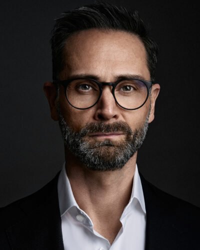 Ein Mann mit Brille und Bart, bekleidet mit einem weißen Hemd und einer dunklen Jacke, blickt vor einem dunklen Hintergrund direkt in die Kamera und schafft so ein professionelles Businessfoto. Sebastian Schueler – Business Fotograf Frankfurt am Main Business Portrait Fotograf Frankfurt Sebastian Schueler Foto 33
