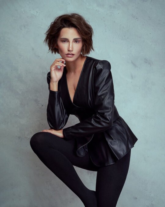 Eine Person mit kurzen Haaren, einer schwarzen Lederjacke und schwarzen Leggings posiert selbstbewusst vor einem grau strukturierten Hintergrund und strahlt eine moderne, professionelle Ausstrahlung aus, die perfekt für ein kommerzielles Shooting ist. Sebastian Schueler – Business Fotograf Frankfurt am Main Model Shooting Janina Schiedlofsky Frankfurt Sebastian Schueler Foto 4