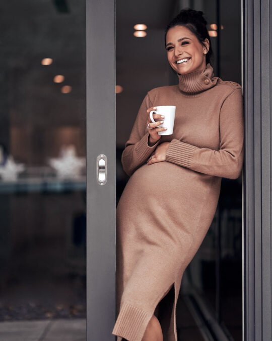 Eine Frau steht in einer Tür, hält eine Tasse, trägt ein langes braunes Pulloverkleid und lächelt. Damit fängt sie die perfekte Essenz der Lifestyle-Fotografie ein. Sebastian Schueler – Business Fotograf Frankfurt am Main Portrait Fotograf Frankfurt Amira Sebastian Schueler Foto