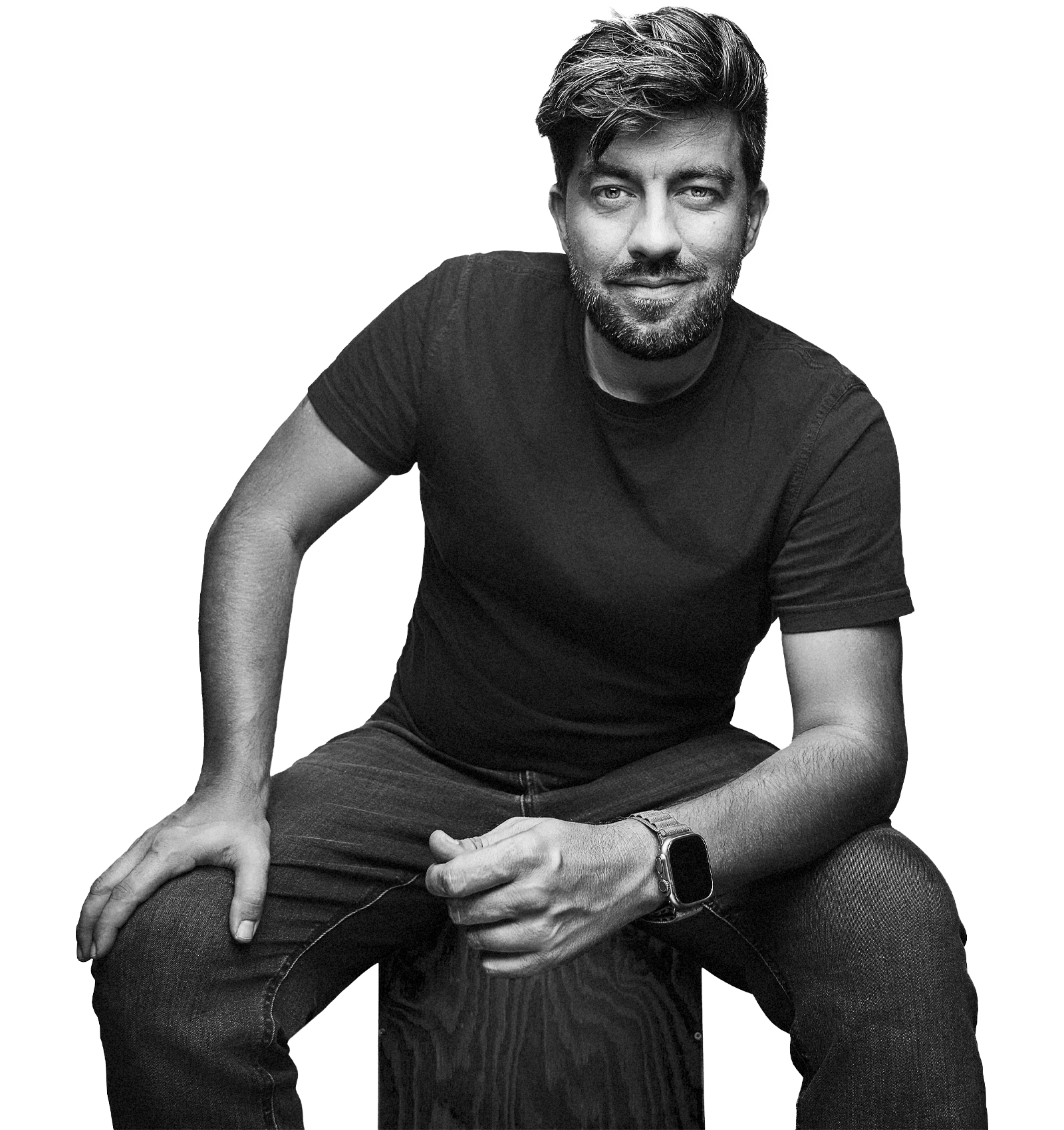 Ein Mann in schwarzem T-Shirt und Jeans sitzt auf einem Hocker, nach vorne gebeugt, eine Hand ruht auf seinem Knie, ein Arm ruht auf dem anderen Knie. Er hat einen gestutzten Bart und lächelt leicht. Sebastian Schueler - Business Fotograf Frankfurt am Main