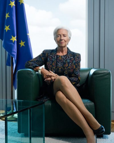 Eine Frau mit kurzen weißen Haaren sitzt auf einem dunkelgrünen Sessel mit gekreuzten Beinen in der Nähe einer EU-Flagge in einer modernen Büroumgebung. Sebastian Schueler – Business Fotograf Frankfurt am Main Christine Lagarde EZB Business Portrait Fotograf Frankfurt Sebastian Schueler