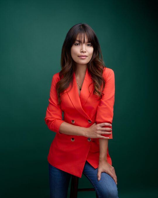 Eine Frau mit langen braunen Haaren, die einen leuchtend orangefarbenen Blazer und blaue Jeans trägt, sitzt auf einem Hocker vor einem grünen Hintergrund. Sebastian Schueler – Business Fotograf Frankfurt am Main Business Portrait Fotograf Frankfurt Sebastian Schueler WEB without Grain