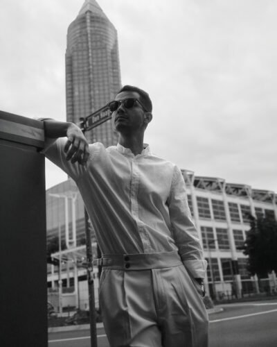 Ein Mann mit Sonnenbrille und weißem Hemd lehnt sich an ein Gebäude in der Nähe eines Straßenschildes, mit einem hohen Gebäude und moderner Architektur im Hintergrund. Sebastian Schueler – Business Fotograf Frankfurt am Main Business Portrait Fotograf Frankfurt Sebastian Schueler WEB DSF4669